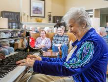 A smiling woman playing the piano for a group of residents.